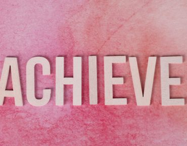 The word 'Achieve' in wooden letters on a textured pink background, symbolizing success and motivation.